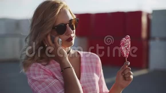 美丽的年轻女子在街上使用智能手机舔着彩色棒棒糖视频的预览图
