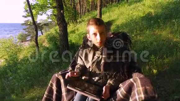 少年坐在一个大湖岸的椅子上青少年男孩热衷于在平板电脑上玩电脑游戏视频的预览图