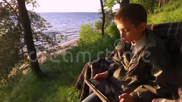 少年坐在一个大湖岸的椅子上青少年男孩热衷于在平板电脑上玩电脑游戏视频的预览图