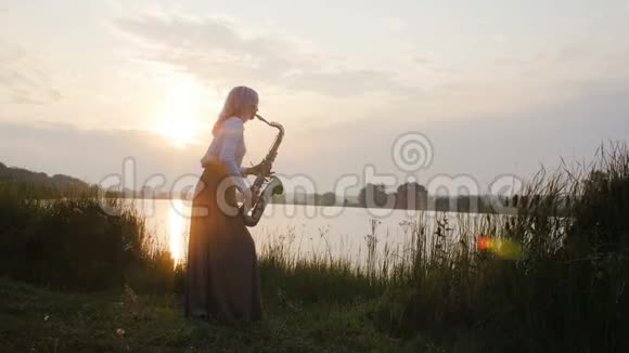 黎明时分一位年轻女子在河岸附近的萨克斯管上演奏一位穿着一件长带着风的美丽女孩的剪影视频的预览图