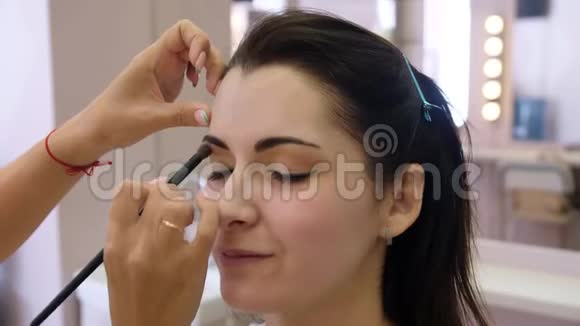 双手化妆师将化妆涂在一个年轻女人的脸上在专业沙龙里做眼妆的女孩晚上视频的预览图