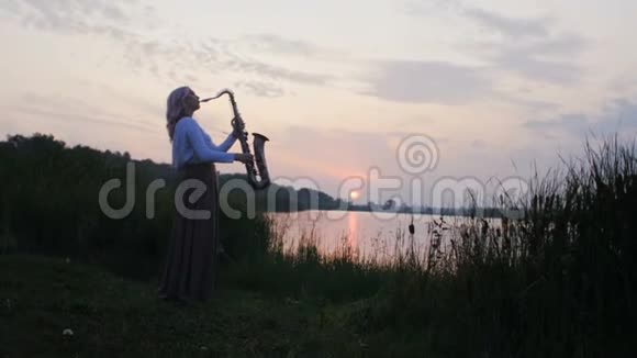 黎明时分一位年轻女子在河岸附近的萨克斯管上演奏一位穿着一件长带着风的美丽女孩的剪影视频的预览图