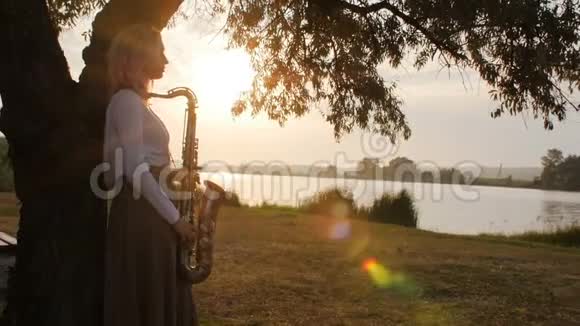 黎明时分一位年轻女子在河岸附近的萨克斯管上演奏一位穿着一件长带着风的美丽女孩的剪影视频的预览图