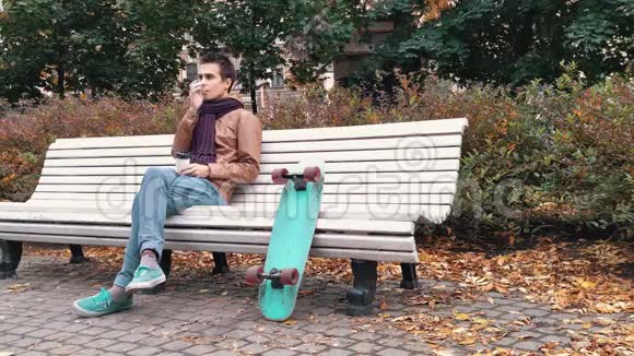 秋天公园里坐在长椅上的带着滑板的针织围巾的年轻人用纸杯喝咖啡视频的预览图