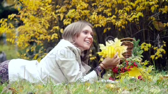 快乐的年轻女子躺在公园里的柳条篮子附近的秋叶上手里拿着一串叶子4k视频的预览图
