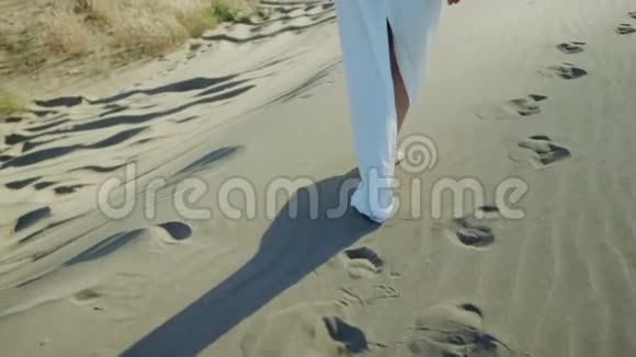 穿着白色连衣裙在沙滩沙丘上行走的女人的脚太阳落山的影子视频的预览图