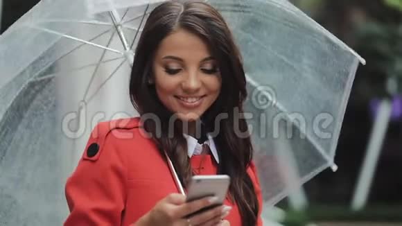 美丽的年轻商务女性在雨天的街道上使用智能手机微笑拿着雨伞交流视频的预览图