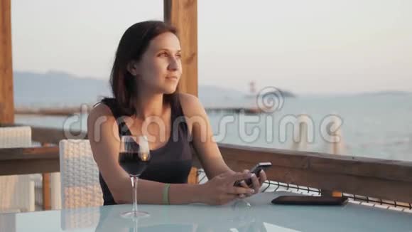 年轻的布鲁内特在日落时坐在海边的咖啡馆里用她的智能手机端着一杯红酒视频的预览图