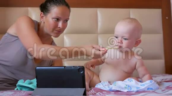 年轻的妈妈用一勺粥喂可爱的宝宝宝宝坐在床上盯着平板电脑上的卡通片视频的预览图