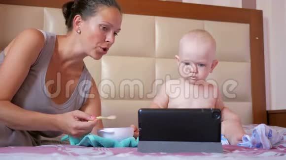 年轻的妈妈用一勺粥喂可爱的宝宝宝宝坐在床上盯着平板电脑上的卡通片视频的预览图