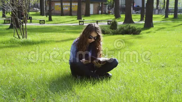 美丽的年轻女子坐在草地上的公园里看书视频的预览图