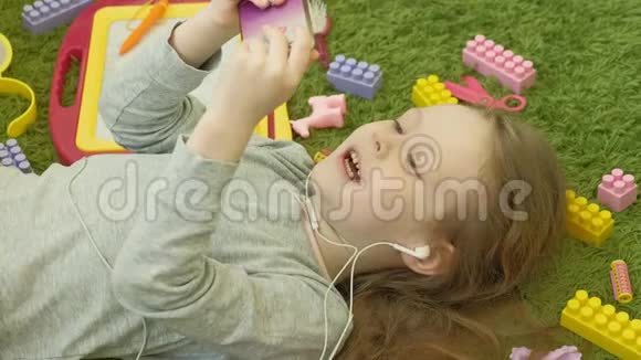 小女孩躺在绿色背景耳机和使用电话顶部视图视频的预览图