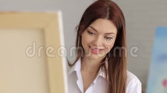一个穿着白色衬衫的年轻女孩在画室的画布上画画艺术视频的预览图