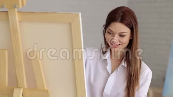 一个穿着白色衬衫的年轻女孩在画室的画布上画画艺术视频的预览图