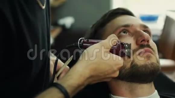 有才华的留胡子的理发师正在理发店里用黑色的剪发披风修剪他的客户的胡须他用的是视频的预览图