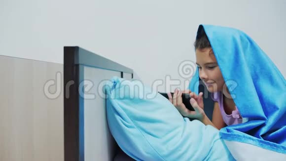 带智能手机的毯子下可爱女孩的肖像小女孩在社交媒体毯子下玩网络游戏女孩视频的预览图