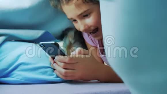 带智能手机的毯子下可爱女孩的肖像在社交媒体下玩网络游戏的小女孩的生活方式视频的预览图