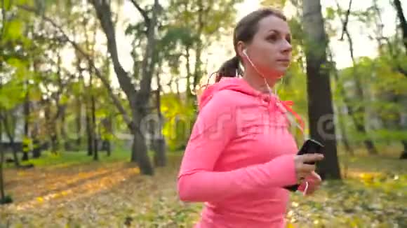 日落时分一位戴着耳机和智能手机的女士在秋天的公园里奔跑慢动作视频的预览图
