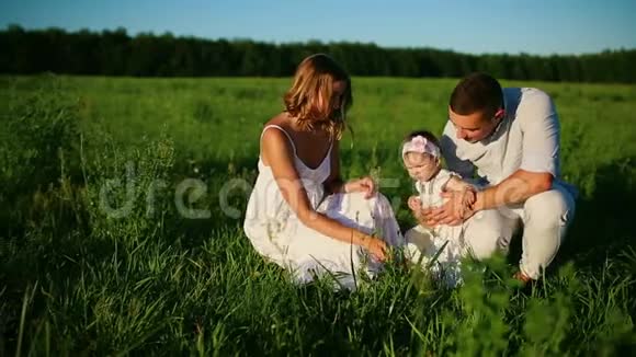 家庭幸福爸爸妈妈和一个小女孩在夕阳下穿着白色衣服的田野里散步视频的预览图