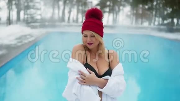 年轻的金发女郎穿着浴袍在冬天的室外游泳池附近有一间红色的小屋视频的预览图