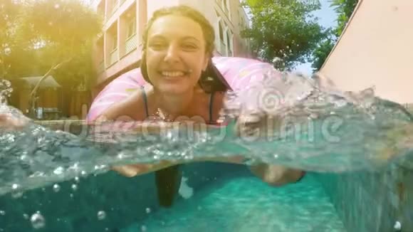 阳光明媚的夏日戴着蓝色比基尼太阳镜的女人躺在充气的粉红色甜甜圈里漂浮在游泳池里看镜头视频的预览图