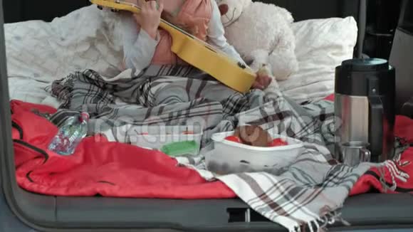 美丽的小女孩坐在一辆敞篷的汽车后备箱里在河岸上玩黄色的四弦琴视频的预览图