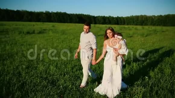 家庭幸福爸爸妈妈和一个小女孩在夕阳下穿着白色衣服的田野里散步视频的预览图