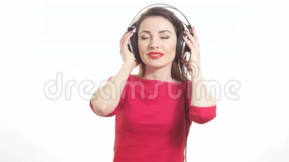 可爱的红衣女郎听着音乐摘下大耳机对着镜头眨眨眼在白色背景上微笑着视频的预览图
