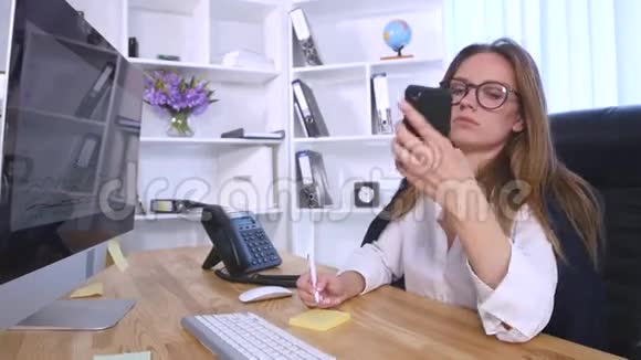 年轻的商务女性坐在办公室的桌子旁一边用手机说话一边在笔记本上做笔记视频的预览图