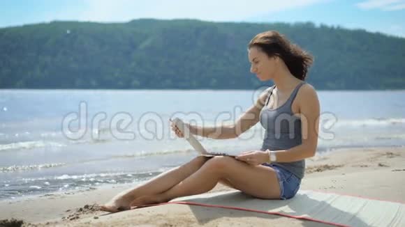 一位美丽的年轻女士正在用笔记本电脑坐在海滩上视频的预览图