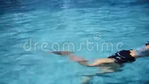 穿着泳衣的女人漂浮在蓝色的游泳池里放松用她的腿溅起水花视频的预览图
