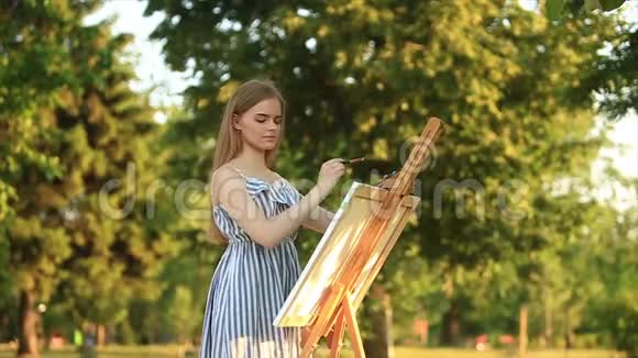 美丽的女孩站在公园里用颜料和铲子画一幅画视频的预览图