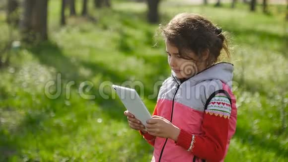 在森林里绿草如茵的背景下春天带着平板电脑的女孩在玩网络游戏视频的预览图