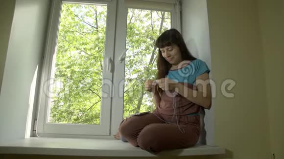年轻女子用坐在窗台上的针织针编织蓝色的纱线视频的预览图