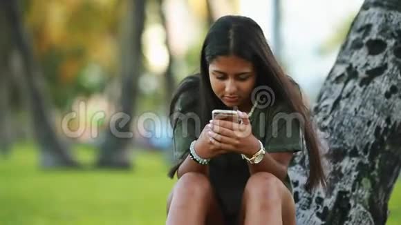 少女坐在一棵树旁用手机视频的预览图