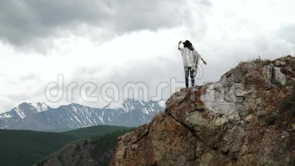 年轻漂亮的女旅行者戴着帽子和斗篷在山顶上放松群山环绕视频的预览图