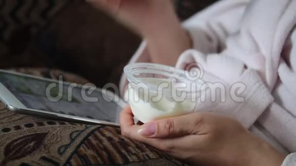一个穿着家庭浴袍的女孩躺在沙发上用平板电脑工作同时他吃酸奶视频的预览图