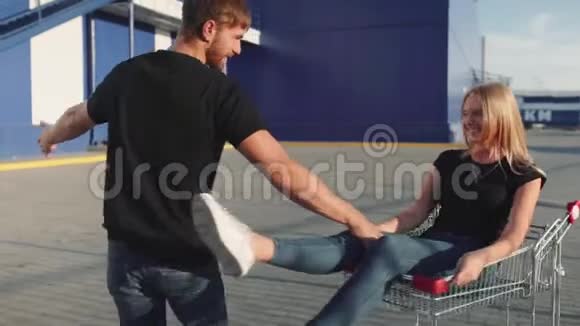 日落时分这家伙在超市停车场的手推车里翻滚着迷人的年轻女人慢动作视频的预览图