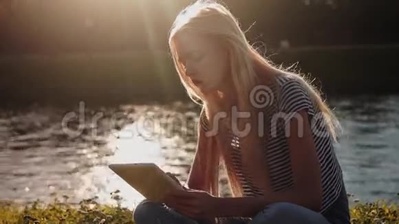 坐在河边绿草如茵的平板电脑上的小女孩视频的预览图