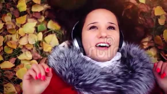美丽幸福的年轻女子戴着耳机躺在秋叶上听音乐视频的预览图
