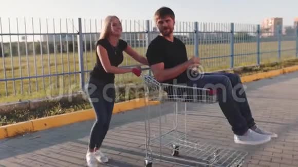 日落时分年轻的女人在超市停车场的手推车里翻滚着迷人的家伙慢动作视频的预览图
