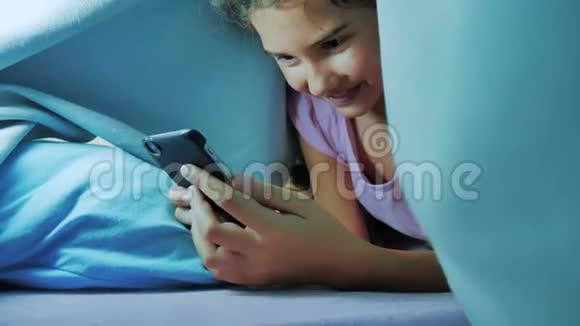 带智能手机的毯子下可爱女孩的肖像生活方式下玩网络游戏的小女孩社交媒体视频的预览图