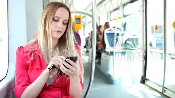 年轻的金发女人骑着电车在手机手机手机上打字拿着眼镜视频的预览图