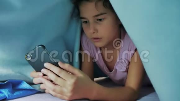 带智能手机的毯子下可爱女孩的肖像在社交媒体下玩生活方式网络游戏的小女孩视频的预览图