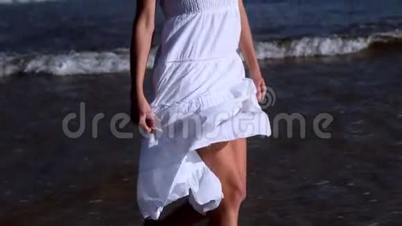 阳光明媚的一天美丽的金发女郎穿着白色连衣裙在海滩上散步视频的预览图