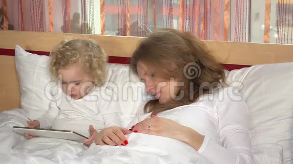 母亲和蹒跚学步的女儿带着平板电脑躺在床上视频的预览图