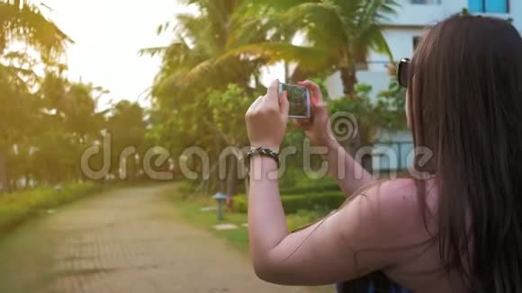 漂亮的快乐女孩戴着眼镜和一件蓝色的连衣裙正在手机上拍摄夕阳的照片绿色的手掌和视频的预览图