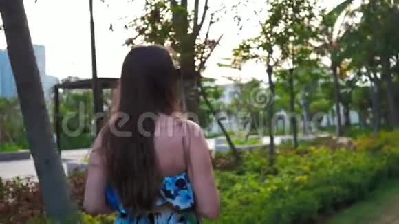 漂亮的快乐女孩戴着眼镜和一件蓝色的连衣裙正在手机上拍摄夕阳的照片绿色的手掌和视频的预览图
