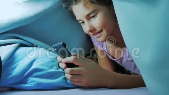 带智能手机的毯子下可爱女孩的肖像在生活方式社交媒体下玩网络游戏的小女孩视频的预览图