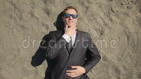 一个穿西装戴太阳镜的商人躺在冲浪线附近的海滩上视频的预览图